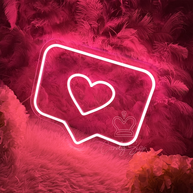 May include: A white neon sign shaped like an Instagram comment bubble with a heart inside. The sign is on a pink fluffy background.