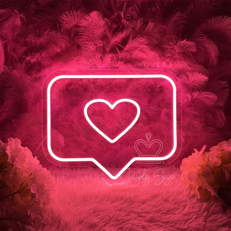 May include: Pink neon sign shaped like an Instagram like button with a white heart inside. The sign is on a pink fluffy background.