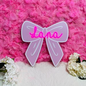 May include: A pink bow-shaped sign with the name "Lana" in a cursive font. The sign is set against a background of pink feathers. White flowers are visible at the bottom left and right corners of the image.