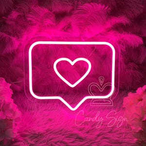 May include: Pink neon sign shaped like a speech bubble with a white heart inside. The sign is on a pink fluffy background.