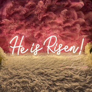 May include: White neon sign with the words "He is Risen!" against a background of pink feathers.