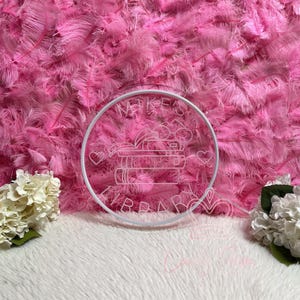 May include: A white circular neon sign with the text "Mike's Library" and a stack of books, hearts, and other decorative elements. The sign is set against a backdrop of pink feathers and white faux fur.