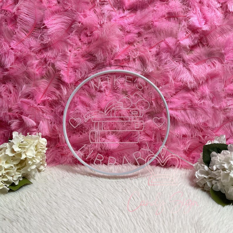 May include: A white circular neon sign with the text "Mike's Library" and a stack of books, hearts, and other decorative elements. The sign is set against a backdrop of pink feathers and white faux fur.
