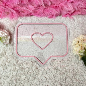 May include: Pink neon sign shaped like a speech bubble with a heart inside. The sign is on a white fluffy surface.