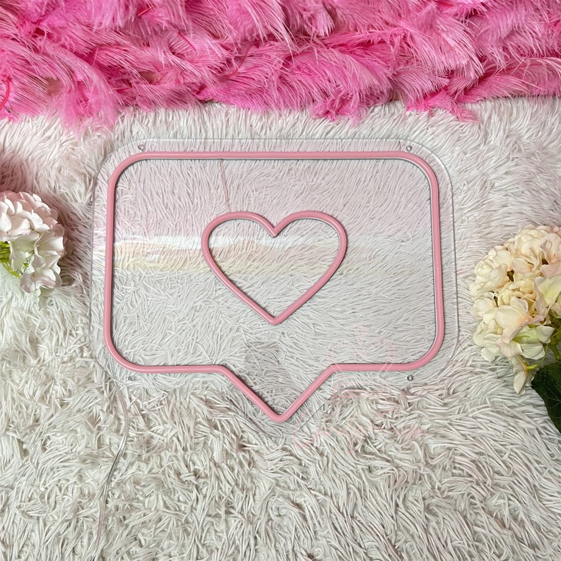 May include: Pink neon sign shaped like a speech bubble with a heart inside. The sign is on a white fluffy surface.