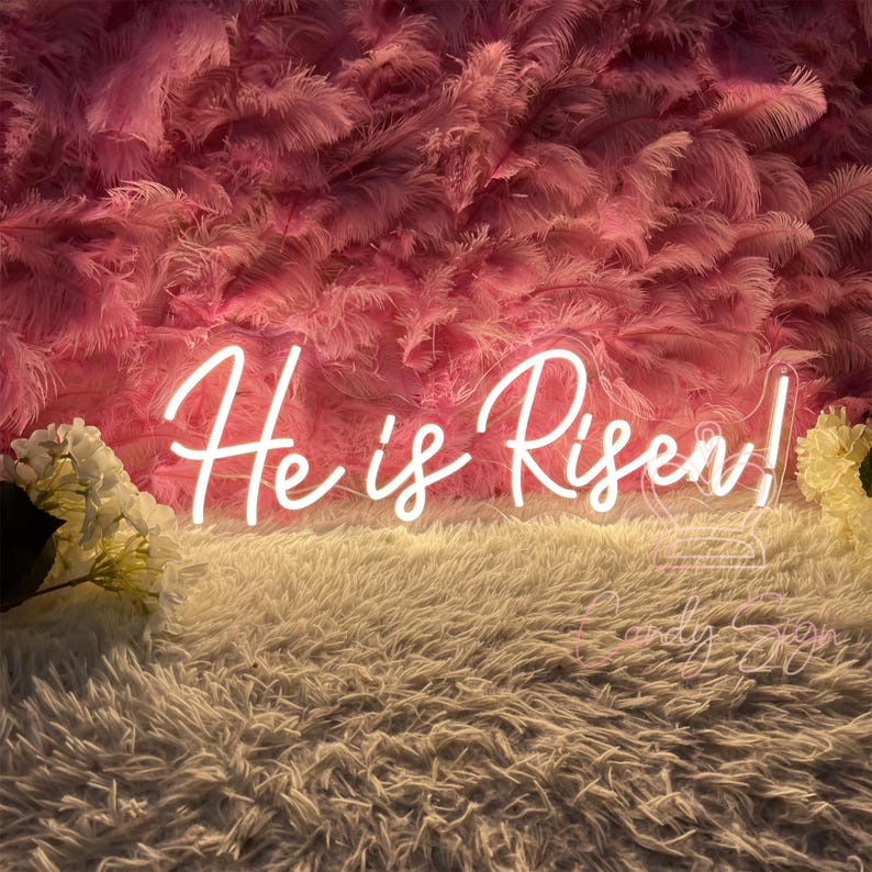 May include: White neon sign with the text "He is Risen!" against a pink feather backdrop.