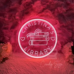 May include: A neon sign with the text "CHRISTINA'S LIBRARY" in a circular design. The sign features a stack of books with a ribbon, hearts, and the words "Candy Sign" at the bottom. The sign glows with a pink light.