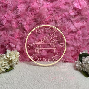 May include: A neon sign with the text "Mike's Library" in a circular frame. The sign features a stack of books, glasses, and hearts, set against a pink feather background and white fur.