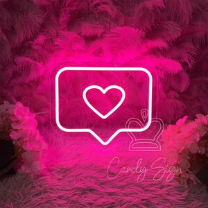 May include: White neon sign shaped like a speech bubble with a heart inside. The sign is lit up in pink and is sitting on a pink fluffy surface.