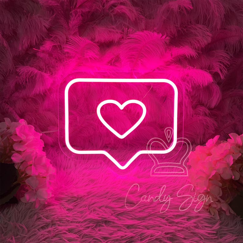 May include: White neon sign shaped like a speech bubble with a heart inside. The sign is lit up in pink and is sitting on a pink fluffy surface.