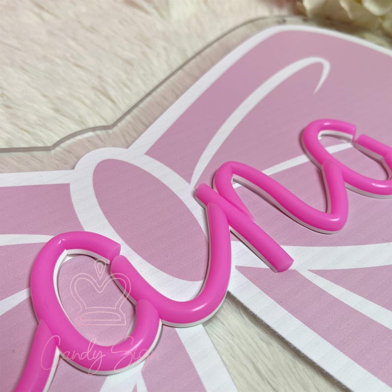May include: A pink and white decorative sign with the word "one" in a cursive font. The sign has a bow-shaped design and the text "Candy Six" is visible.