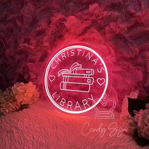 May include: A neon sign with the text "Christina's Library" in a circular design. The sign is white and features a stack of books, hearts, and a ribbon. The background is a fluffy, pink texture with flowers.