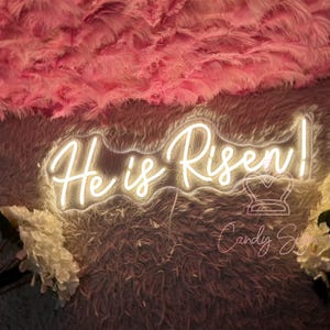 May include: White neon sign with the words "He is Risen!" on a pink fluffy background.