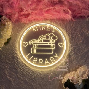 May include: A round, illuminated sign with the words "MIKE'S LIBRARY" in a circular design. The sign features a stack of books and two hearts. The sign is surrounded by a fluffy, white and pink background.