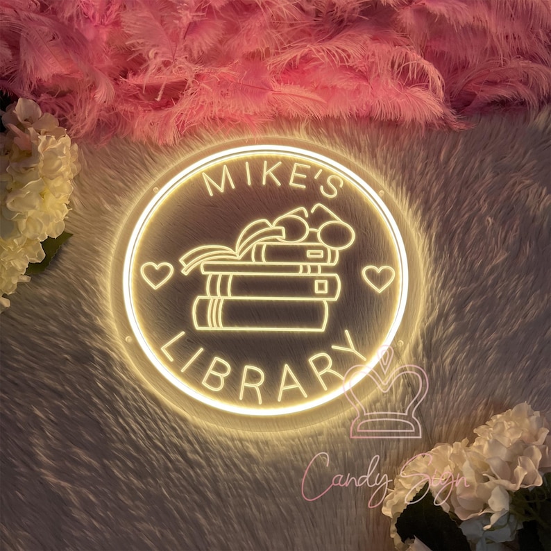 May include: A round, illuminated sign with the words "MIKE'S LIBRARY" in a circular design. The sign features a stack of books and two hearts. The sign is surrounded by a fluffy, white and pink background.
