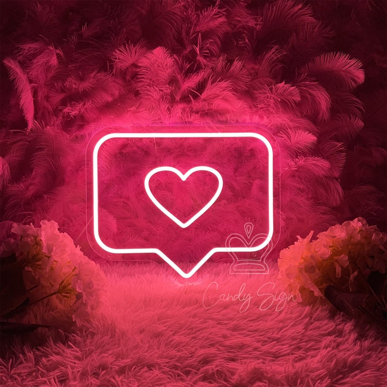 May include: Pink neon sign shaped like a speech bubble with a white heart inside. The sign is on a pink fluffy surface with pink feathers in the background.