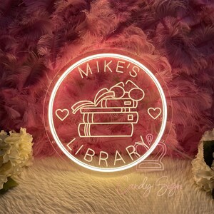 May include: A round, illuminated neon sign with the words "Mike's Library" in a white, cursive font. The sign features a stack of books, glasses, and two hearts. The background is a soft pink with feather-like textures.
