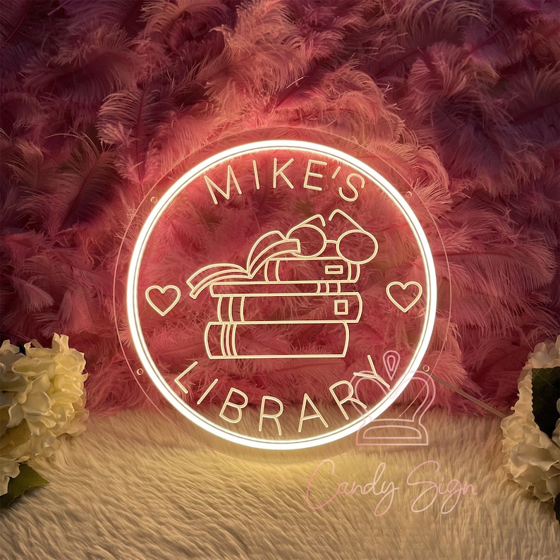 May include: A round, illuminated neon sign with the words "Mike's Library" in a white, cursive font. The sign features a stack of books, glasses, and two hearts. The background is a soft pink with feather-like textures.