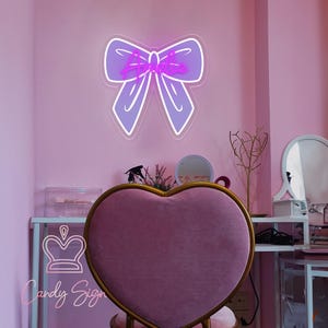 May include: A purple neon bow sign with the word "Ariana" in a cursive font. The sign is mounted on a pink wall. A heart-shaped chair with a gold frame is in the foreground. The room has a vanity area with a mirror and decorative items.