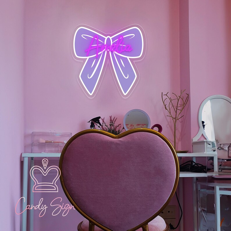 May include: A purple neon bow sign with the word "Ariana" in a cursive font. The sign is mounted on a pink wall. A heart-shaped chair with a gold frame is in the foreground. The room has a vanity area with a mirror and decorative items.