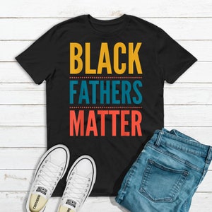May include: Black t-shirt with the text "Black Fathers Matter" in yellow, blue, and red lettering. The shirt is on a white wooden surface with a pair of white sneakers and blue jeans.