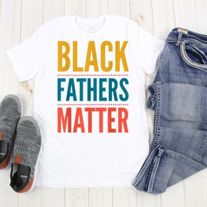 May include: White t-shirt with the text "BLACK FATHERS MATTER" in yellow, teal, and red lettering.