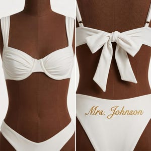 May include: A white bikini set displayed on a mannequin. The top has thick straps and a structured bust. The bottoms are high-waisted with the embroidered text "Mrs. Johnson" in gold. A large bow detail is on the back of the top.