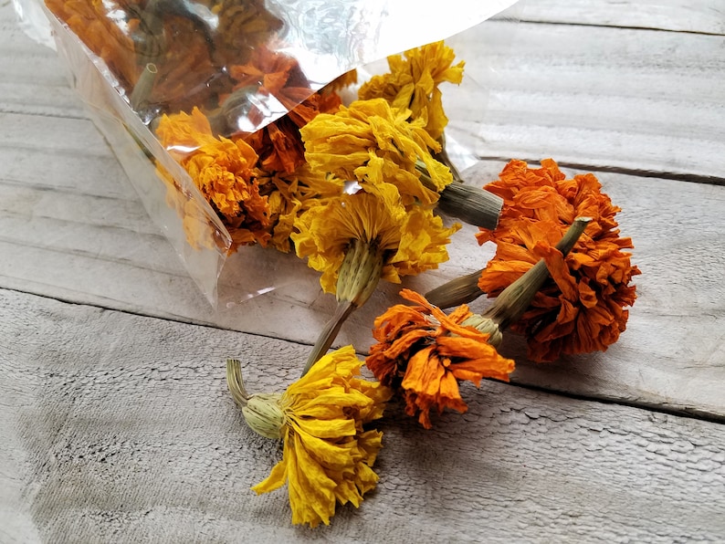 Puede incluir: Flores de cal&eacute;ndula secas en tonos naranja y amarillo, que se derraman de una bolsa de pl&aacute;stico transparente sobre una superficie de madera clara.