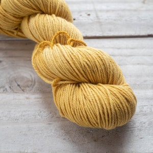 Puede incluir: Un ovillo de hilo amarillo, enrollado en forma de bola, sobre una superficie de madera blanca.