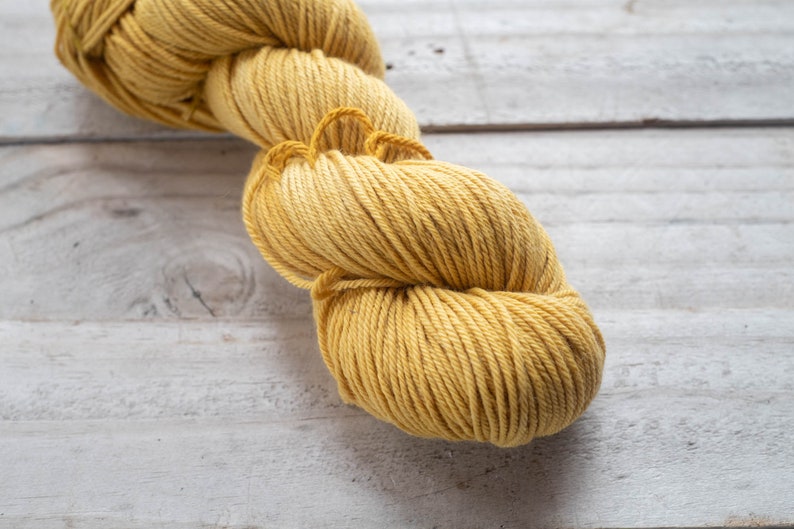 Puede incluir: Un ovillo de hilo amarillo, enrollado en forma de bola, sobre una superficie de madera blanca.