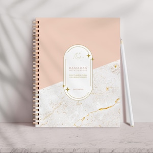 May include: A spiral-bound notebook with a peach and marble design. The cover features gold accents and the text "Ramadan with Purpose". A white pencil rests against the notebook, which is on a light-colored surface.