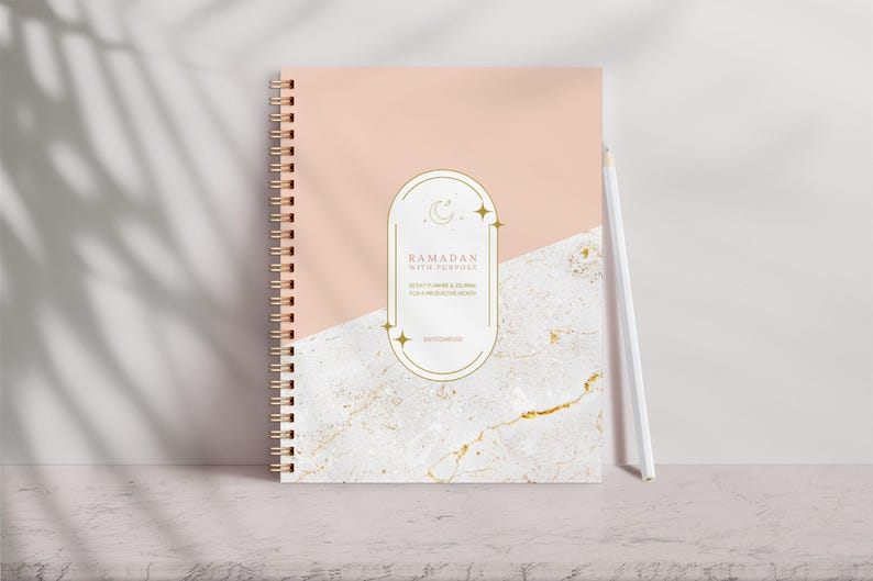 May include: A spiral-bound notebook with a peach and marble design. The cover features gold accents and the text "Ramadan with Purpose". A white pencil rests against the notebook, which is on a light-colored surface.
