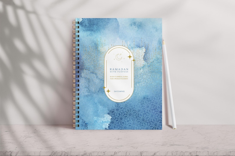 May include: A blue and gold spiral bound journal with the text "Ramadan with Purpose" and a crescent moon and stars design. The journal is on a white surface.