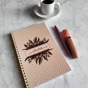 May include: A spiral-bound notebook with a floral design and gold accents, alongside a cup of coffee, a pen, and a lip gloss. The notebook features a geometric pattern and text in Arabic. The items are arranged on a marble surface.