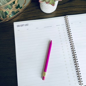 May include: A white spiral bound notebook with the title "Eid Gift List" printed on the cover. The notebook has columns for "For", "Gift", "Price", "Purchased From", "Arrived", "Given", "For", and "Gift". A pink pencil is lying on the open notebook.