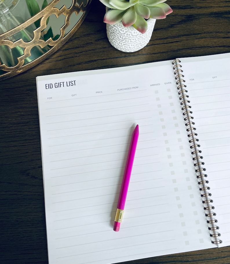 May include: A white spiral bound notebook with the title "Eid Gift List" printed on the cover. The notebook has columns for "For", "Gift", "Price", "Purchased From", "Arrived", "Given", "For", and "Gift". A pink pencil is lying on the open notebook.
