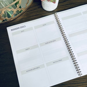 May include: A white planner with a gold spiral binding. The planner has a section for Ramadan goals with headings for Quran, Prayer, Charity, Giving Up Bad Habits, Developing Good Habits, and Health & Fitness.