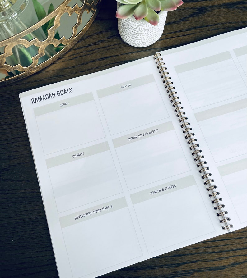 May include: A white planner with a gold spiral binding. The planner has a section for Ramadan goals with headings for Quran, Prayer, Charity, Giving Up Bad Habits, Developing Good Habits, and Health & Fitness.