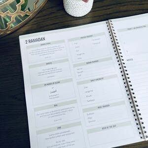 May include: A white and gray planner with a gold spiral binding. The planner has sections for daily tracking of prayer, Quran reading, and other activities. The cover has the text "2 Ramadan" printed on it.