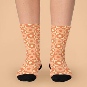 Könnte beinhalten: Ein Paar wadenlange Socken mit einem sich wiederholenden Schachbrettmuster in Orange- und Cremetönen. Jedes Quadrat enthält eine stilisierte Blumengrafik. Die Socken haben einen einfarbigen schwarzen Zehen- und Fersenbereich, der einen deutlichen Kontrast zum gemusterten Stoff bildet, der den Fuß- und Knöchelbereich bedeckt.