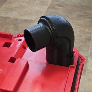 May include: Black plastic dust collection elbow fitting for a power tool. The fitting is attached to a red tool box.