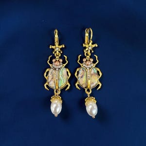May include: A pair of gold-toned dangle earrings featuring a beetle motif. The beetle bodies are inlaid with iridescent abalone shell and clear rhinestones. Each earring is topped with a star-shaped accent and finished with a white pearl drop.