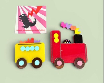 May include: A colorful wooden toy train and a yellow bus with brown wheels. A square card with a pink and white striped background and a black dinosaur illustration is also present.