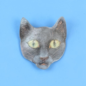 May include: A metal brooch shaped like a cat head with a dark, weathered grey finish and textured surface. The cat features prominent, almond-shaped eyes in a pale, yellowish-green hue, with subtle copper-toned highlights visible on the ears and nose.