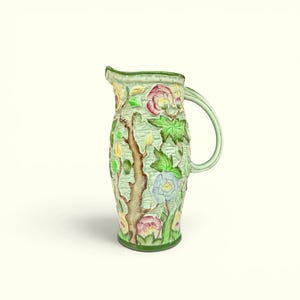 May include: A ceramic jug featuring a textured light green background with a raised relief design of brown tree branches, vibrant green leaves, and multi-coloured flowers in shades of pink, yellow, and light blue, complete with a curved handle.