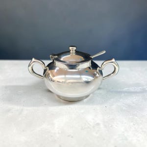 May include: A polished silver-toned sugar bowl with a rounded body, two curved side handles, and a matching lid featuring a small central knob. A small silver spoon rests inside the bowl, with its handle extending through a notch in the lid.