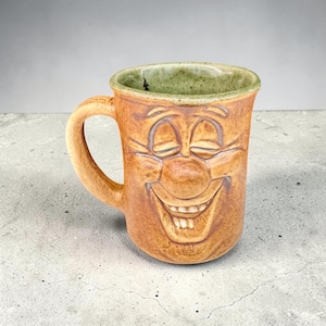 May include: A tan ceramic mug featuring a sculpted, smiling face with closed eyes and visible teeth. The mug has a curved handle on the left side and a textured, light green glazed interior rim, showcasing a handcrafted, rustic appearance.