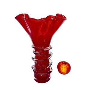 May include: A red glass vase with a wavy, ruffled top and a clear glass spiral design around the base. The vase is sitting on a white surface.