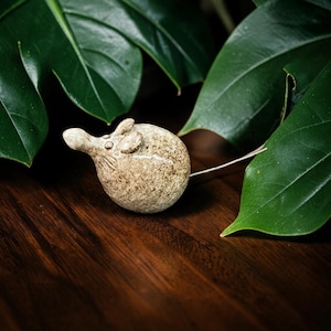 May include: A small, round, ceramic mouse figurine with a speckled beige and brown glaze. The mouse has a smooth, glossy finish, pointed ears, and a thin, metallic wire tail extending from the back of its spherical body.