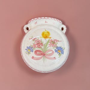 May include: A round ceramic wall pocket with a white glazed finish, featuring a hand-painted floral bouquet in pink, blue, and yellow with a pink ribbon. The piece has a scalloped top edge and two side handles for hanging on a wall surface.